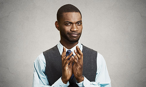 A black man in an vest and button down shirt with tie clasping his hands in front of a gray background.